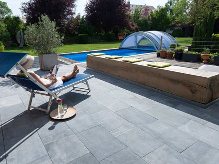 Kvinde slapper af på en liggestol på en terrasse med kalkstensplader, i baggrunden en have med pool