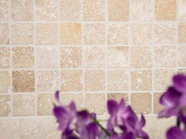 Mosaikfliser i lys beige og fawn veksler i travertinmosaikken Lilla blomster foran en beige-brun travertinmosaik