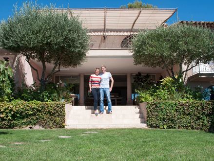 Et par står foran et hotel med travertingulv, en græsplæne i forgrunden og træer til venstre og højre.