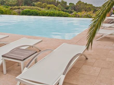 Hvide liggestole på et travertingulv ved en pool, en palmegren stikker ind i billedet fra højre