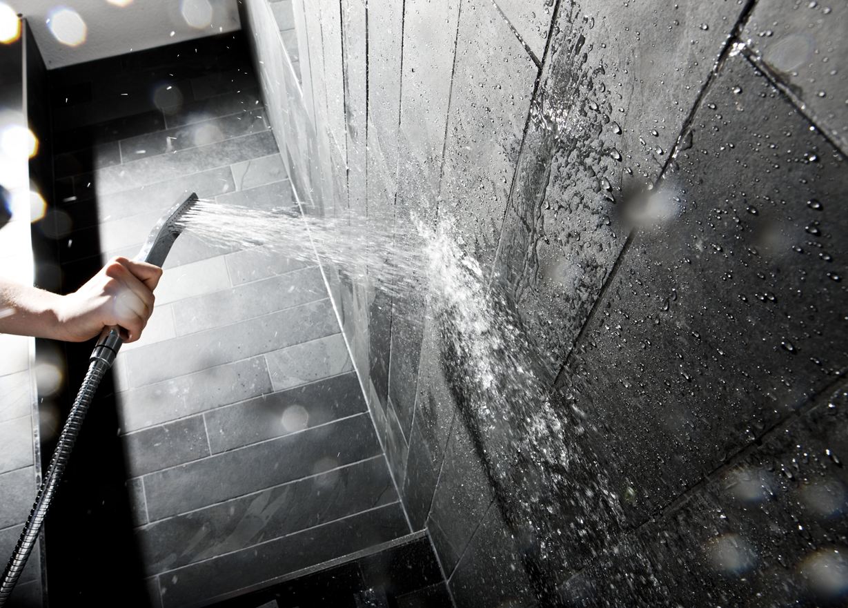 Hånd, der holder et brusehoved mod en væg med mørke stenfliser